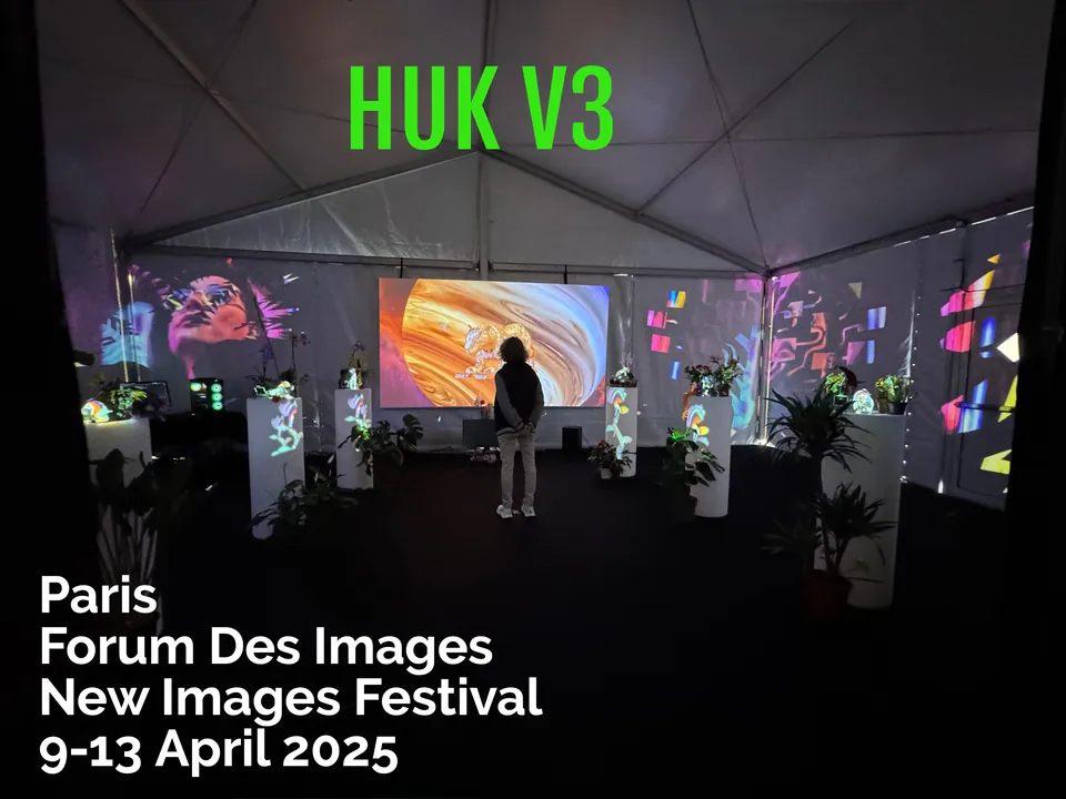 Visitors in a dark tent of projections, flowers and screens. The words Huk V3 above.